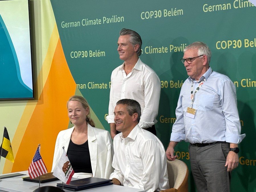 A women and man sit beside each other at a desk with a closed agreement and single pen in the foreground. Flags for California, the US, Germany, and Baden-Wurttemberg sit in the foreground. Two additional leaders stand behind them to pose for a photo. The wallpaper behind them reads, "COP30 Belem, Germany Pavilion."